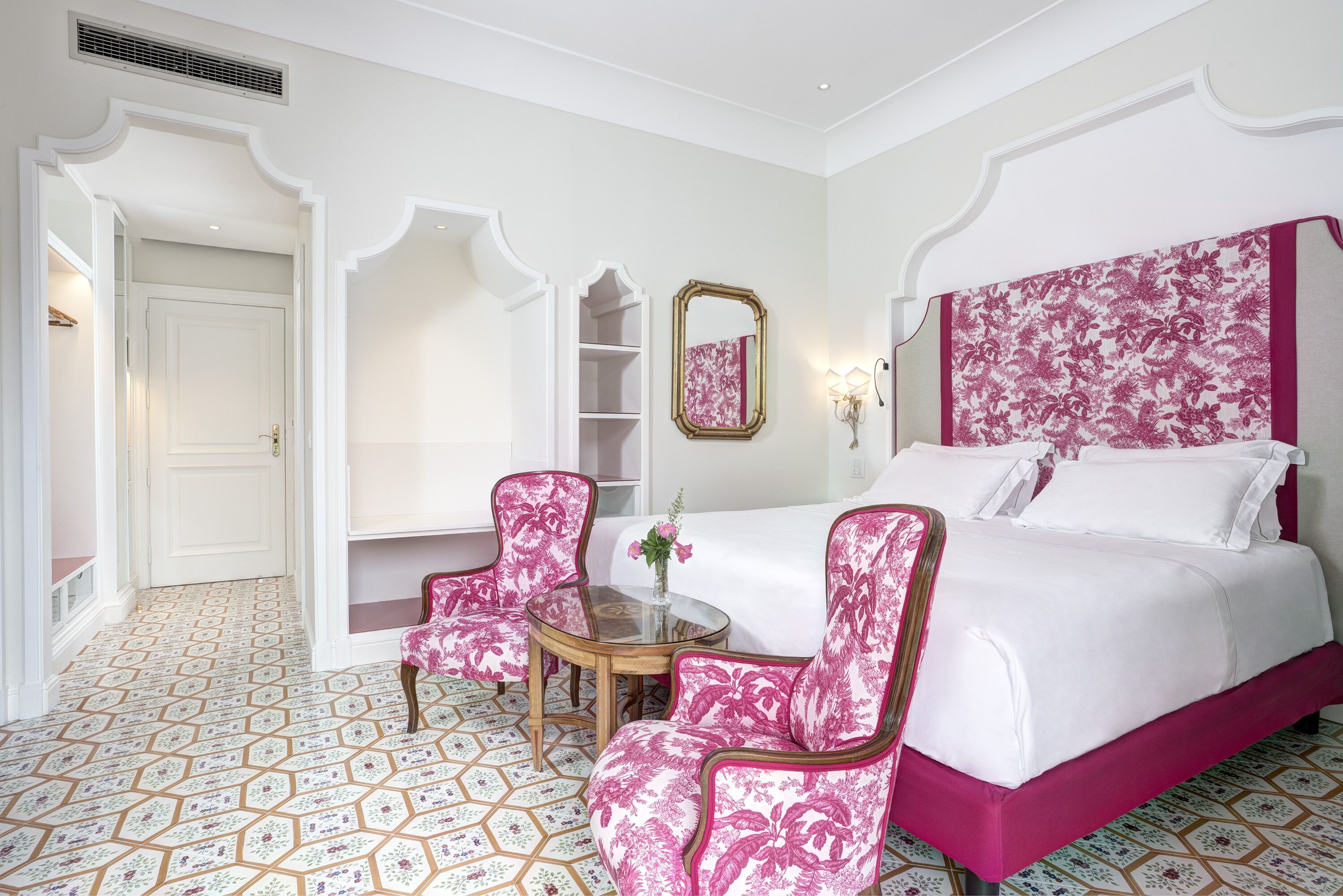 A hotel room with a bed, two pink armchairs, a small table with a vase, and a mirror on the wall.