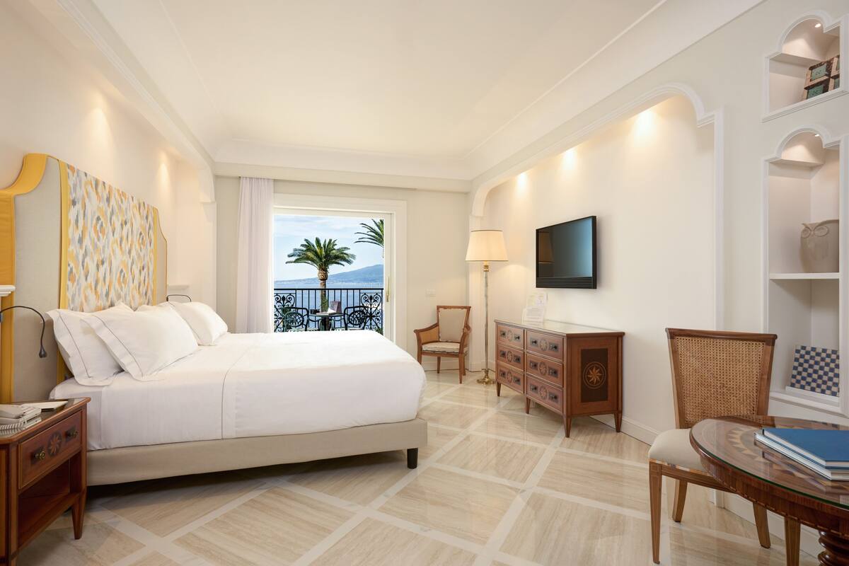 Habitación de hotel con cama, televisión, escritorio y silla. Ventana con vista a palmeras y balcón.