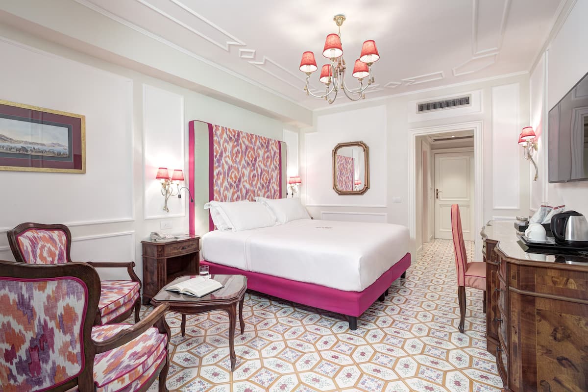Habitación de hotel con una cama grande, un escritorio con silla, una mesita, un espejo y una araña.