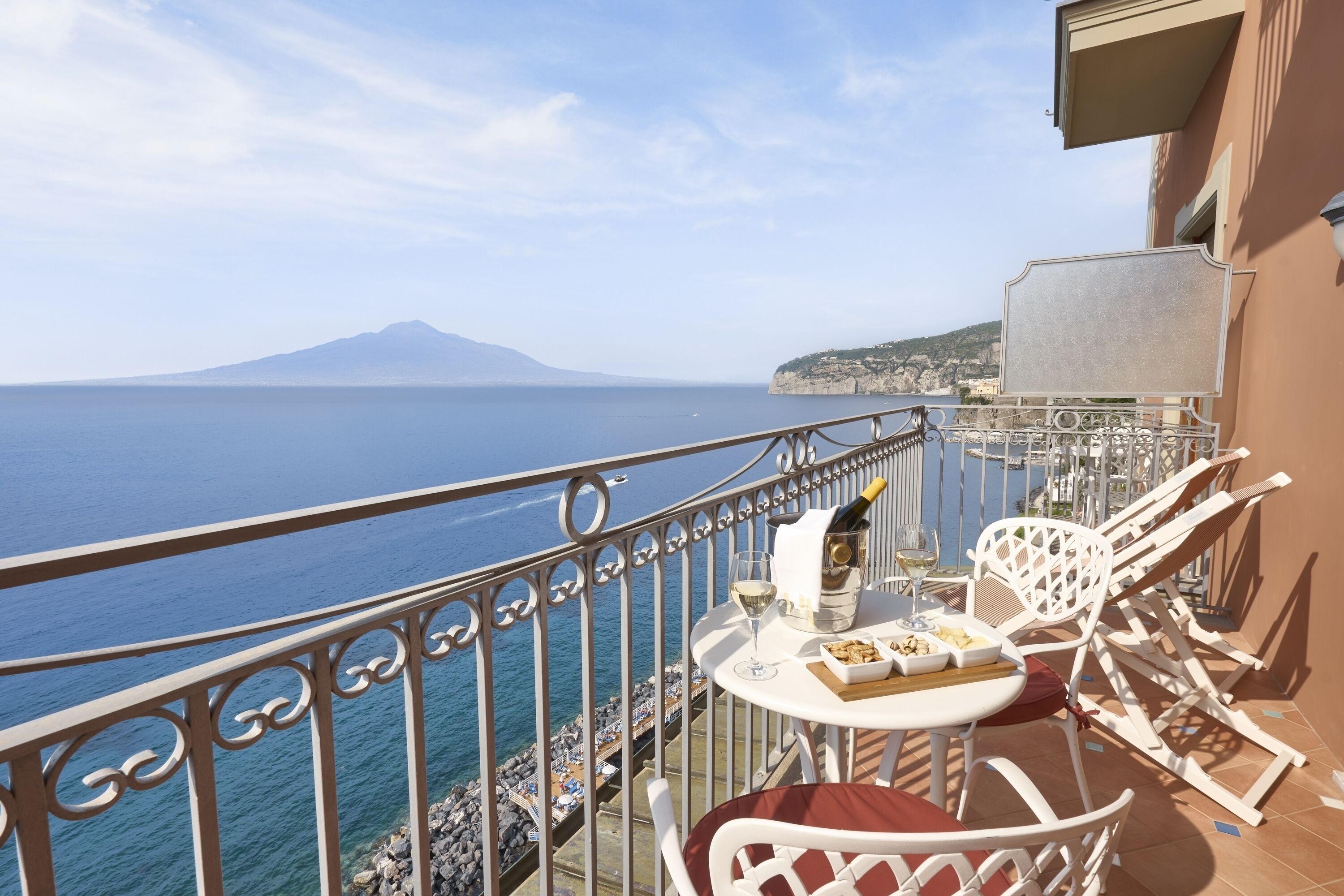 A balcony with a table set for two, overlooking a coastal view with a mountain in the distance.