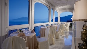 Un restaurant doté de grandes fenêtres en arc qui offrent une vue sur la mer et les montagnes. Les tables sont dressées avec des nappes blanches et des chaises.