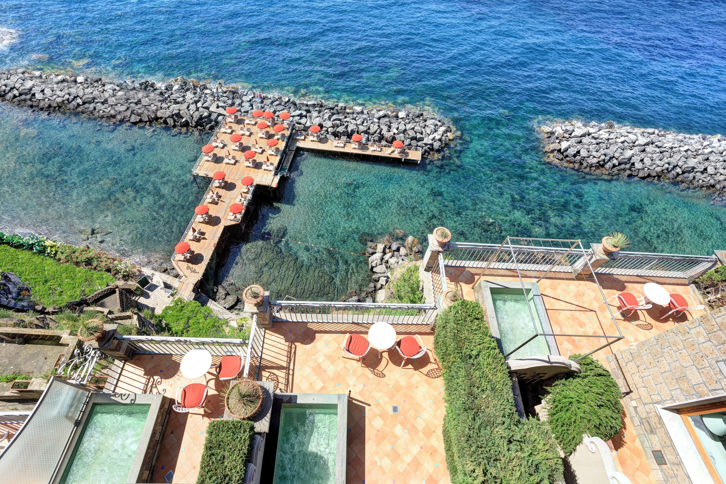 Vista aérea de un resort con una piscina cristalina, zonas de estar al aire libre y un muelle rocoso que se adentra en el agua.