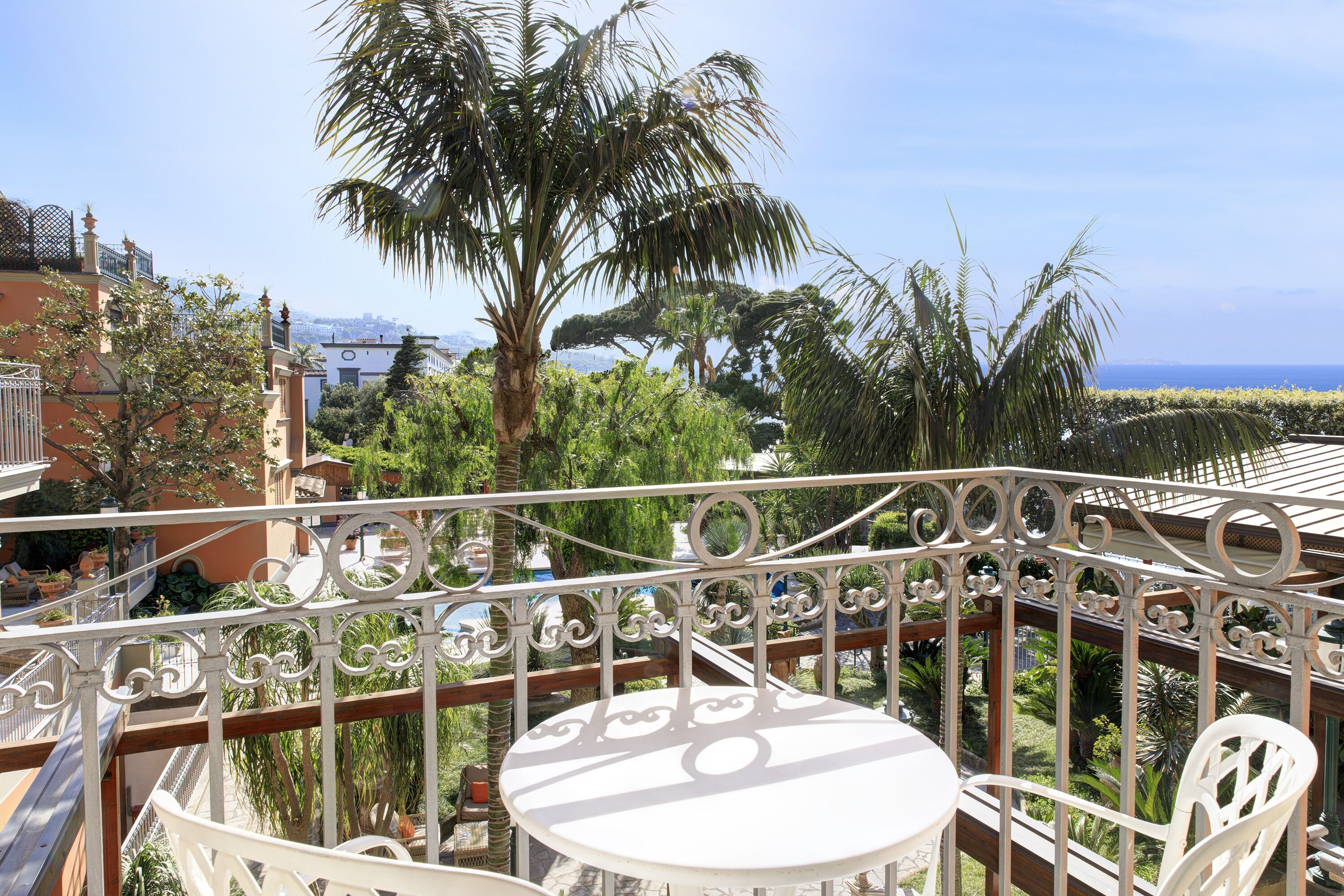 Ein Balkon mit weißem Tisch und Stühlen, umgeben von aufwendig gestalteten Metallgeländern, mit Blick auf einen üppigen Garten und eine ferne Meerlandschaft.