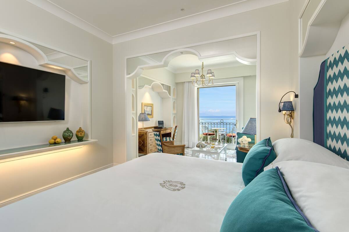 Een hotelkamer met een groot bed, een aan de muur bevestigde televisie, een bureau met stoel, en uitzicht op de zee vanaf een balkon.