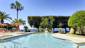 Une piscine entourée de chaises longues et de palmiers, bordée d’une haie bien entretenue et offrant une vue sur la mer.