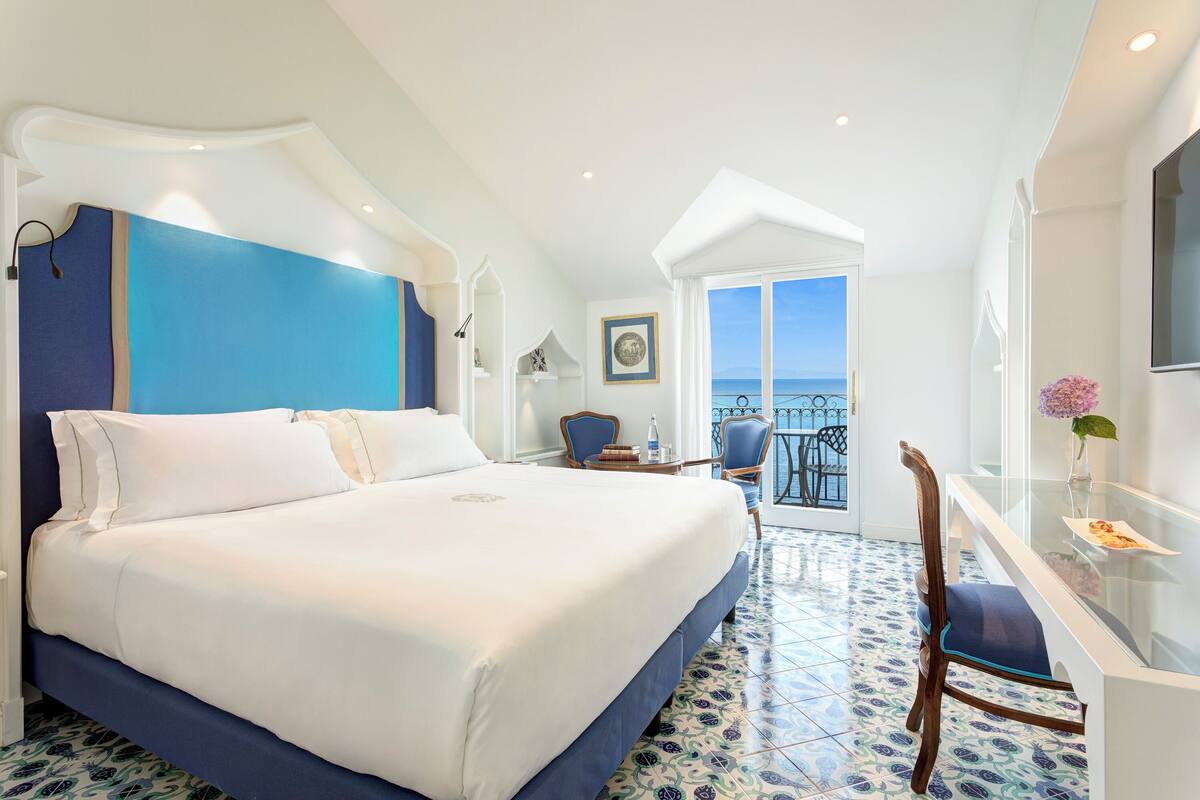 Hotelkamer met een groot bed, een bureau met stoel, een balkon en uitzicht, en een betegelde vloer.