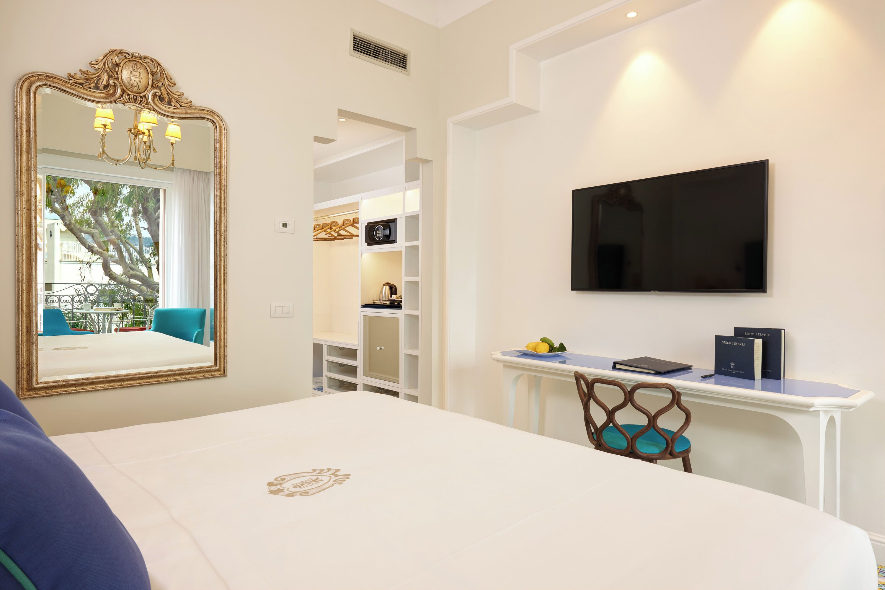 A hotel room with a large bed, a TV mounted on the wall, a desk with a chair, and a mirror with a chandelier.