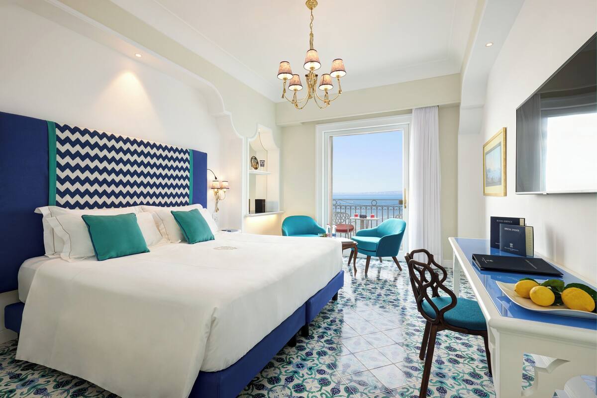 Een hotelkamer met een groot bed, een bureau met een laptop en een fruitschaal, een stoel, en uitzicht op zee.
