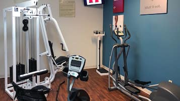 Un gimnasio con máquinas de cardio, equipo de entrenamiento de pesas y una televisión mostrando un programa de ejercicios.