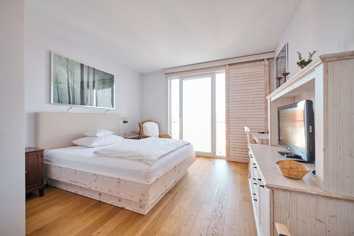 A bedroom with a bed, a television, a window, and a painting on the wall.