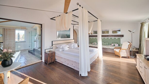 Een slaapkamer met een hemelbed, een houten vloer, een grote spiegel en uitzicht op een groen landschap door een raam.
