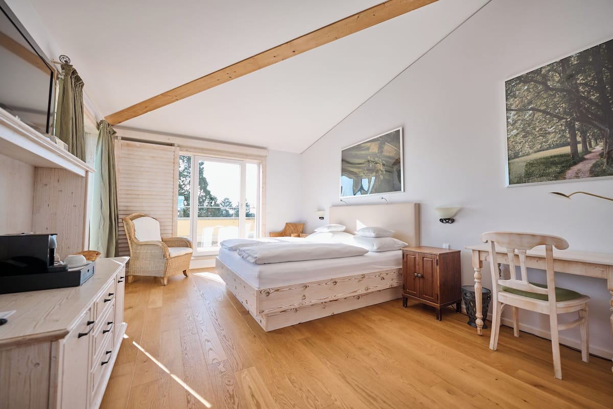 A bedroom with a wooden floor, a bed with white bedding, a desk with a chair, a wicker chair, a cabinet, and a painting on the wall.