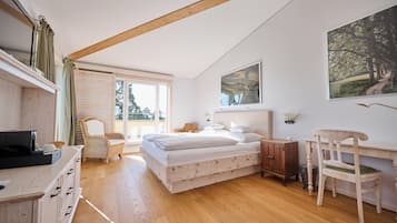 Un dormitorio con piso de madera, una cama con ropa blanca, un escritorio con silla, una silla de mimbre, un armario y un cuadro en la pared.