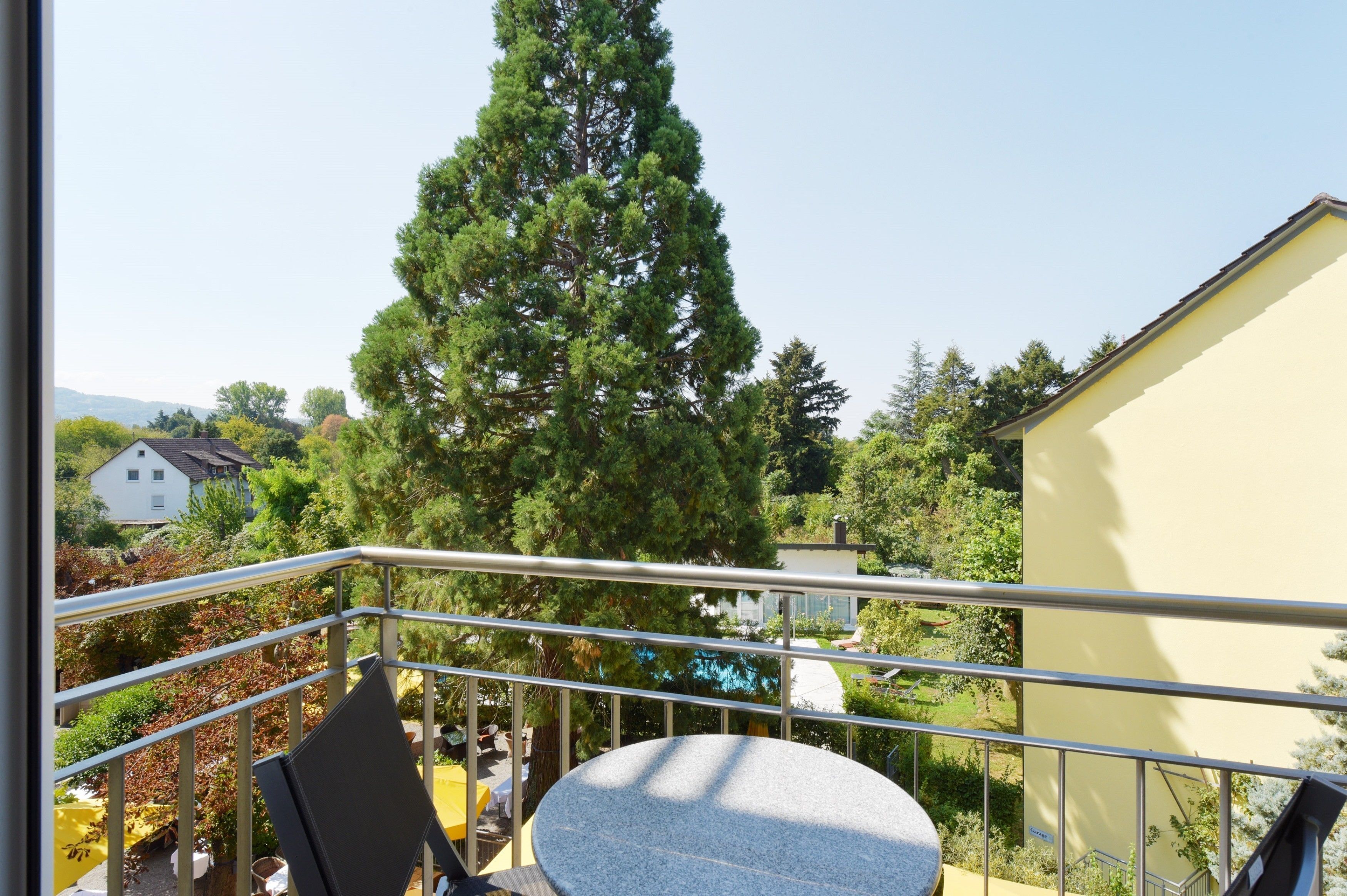 A balcony with a table and chairs, overlooking a garden with a tall tree and a building in the background.