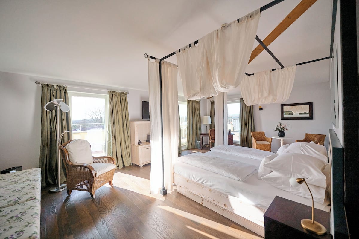 A bedroom with a four-poster bed, a chair, a desk, and a view of the outdoors.