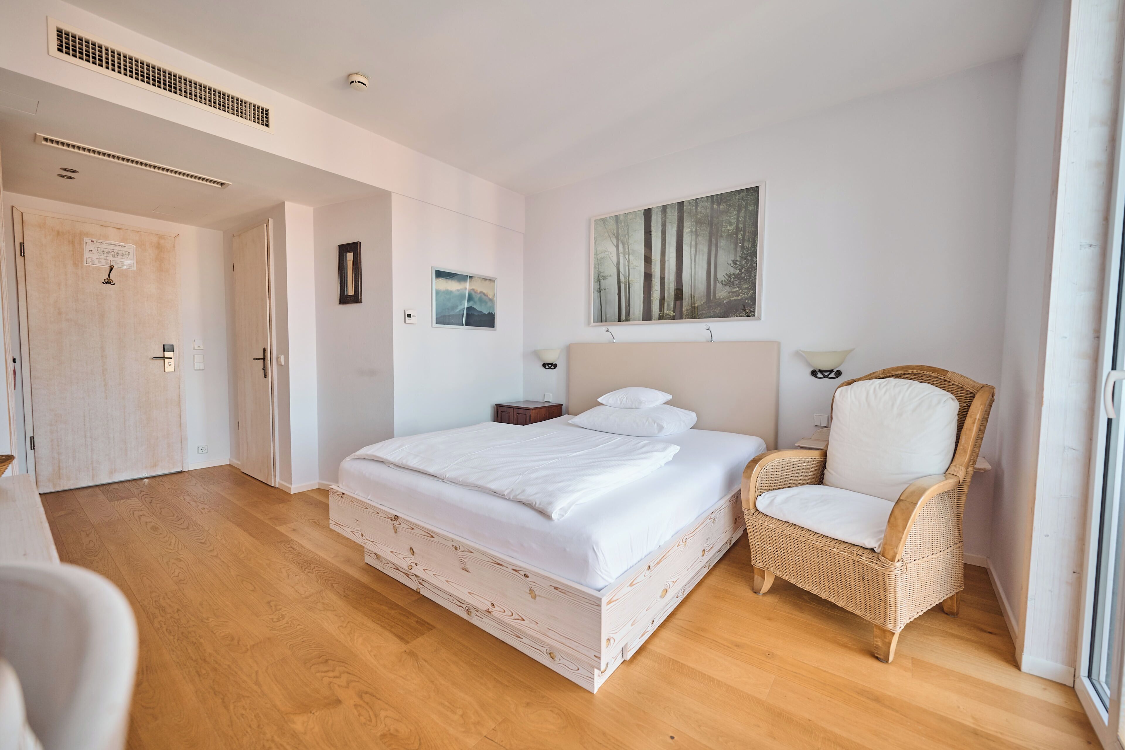 A bedroom with a large bed, a wicker chair, a painting, and a door.