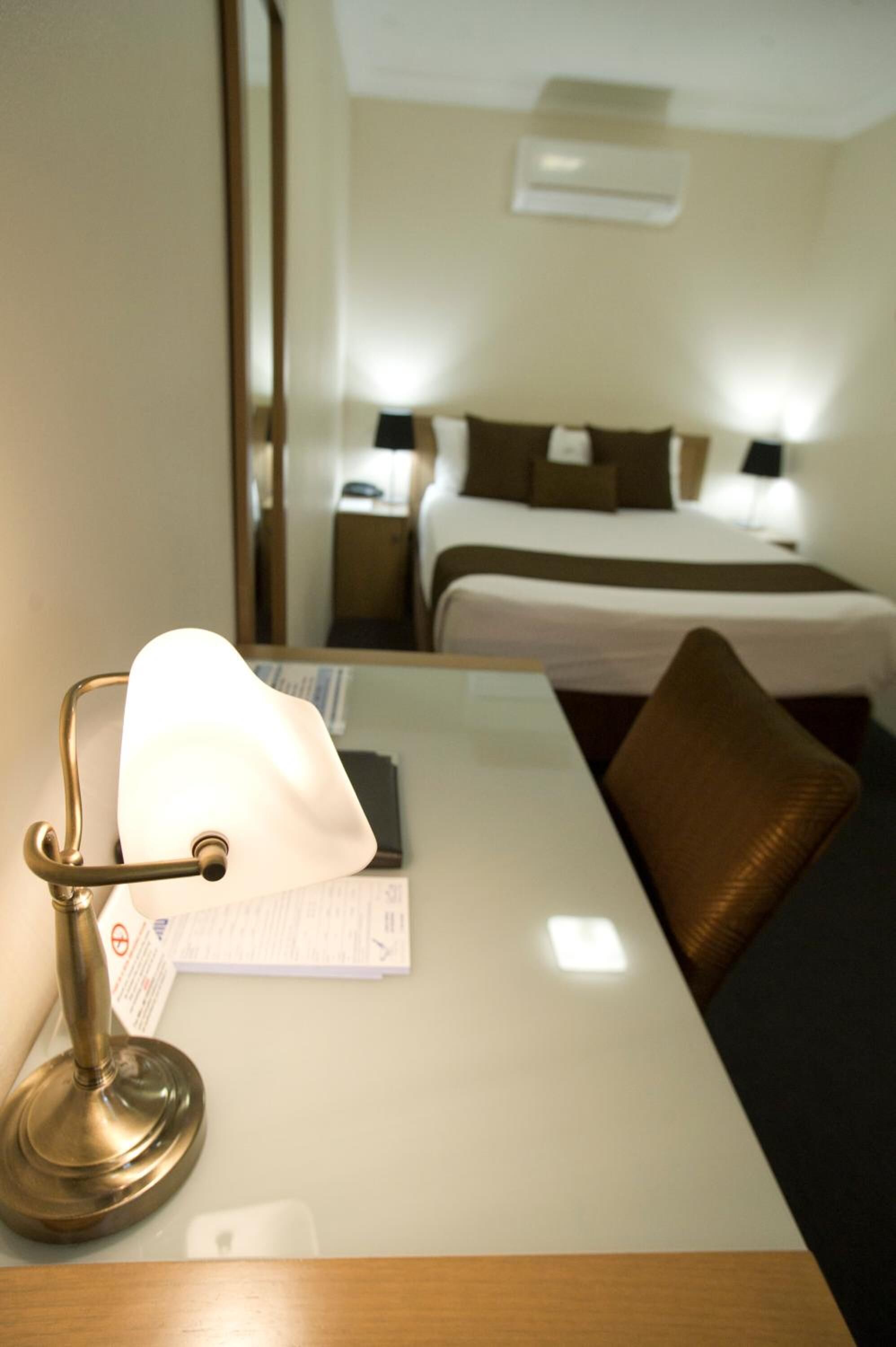 Habitación de hotel con mesita de noche, lámpara, cama con almohadas y un espejo que refleja la habitación.
