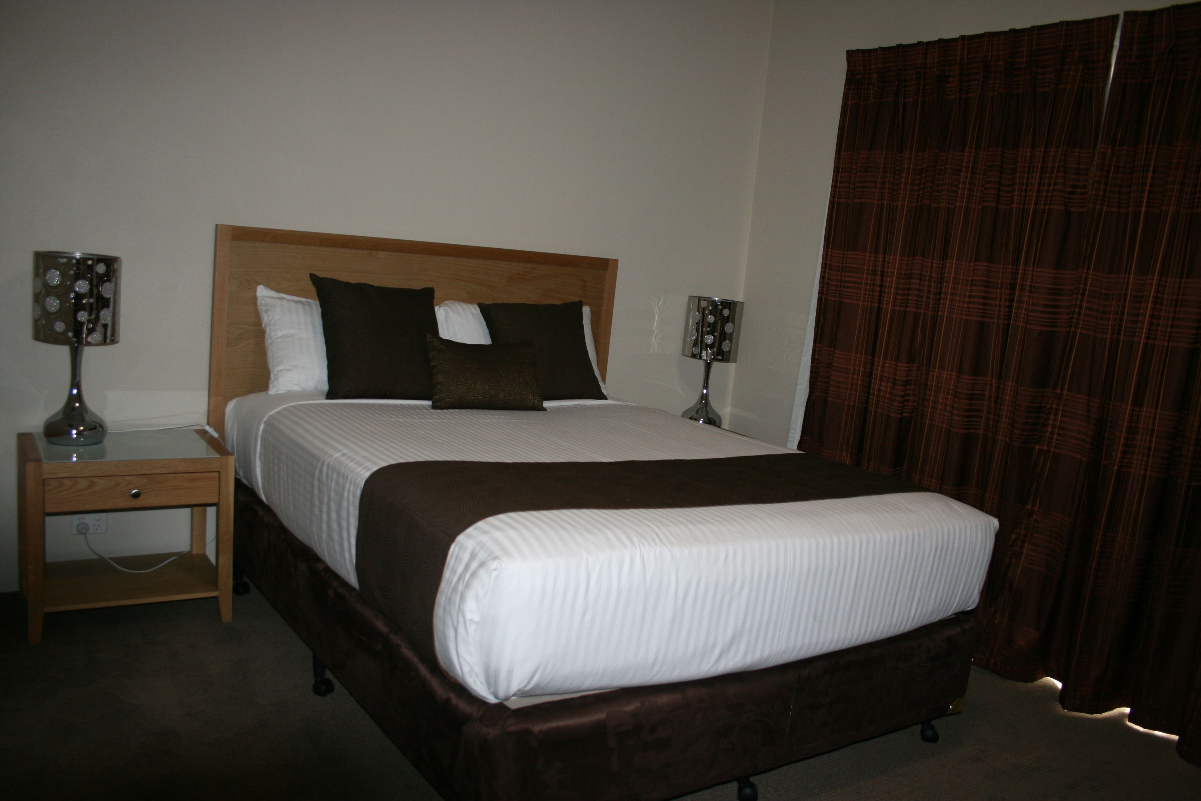 A neatly made bed with a wooden headboard, two bedside lamps, and a brown curtain.