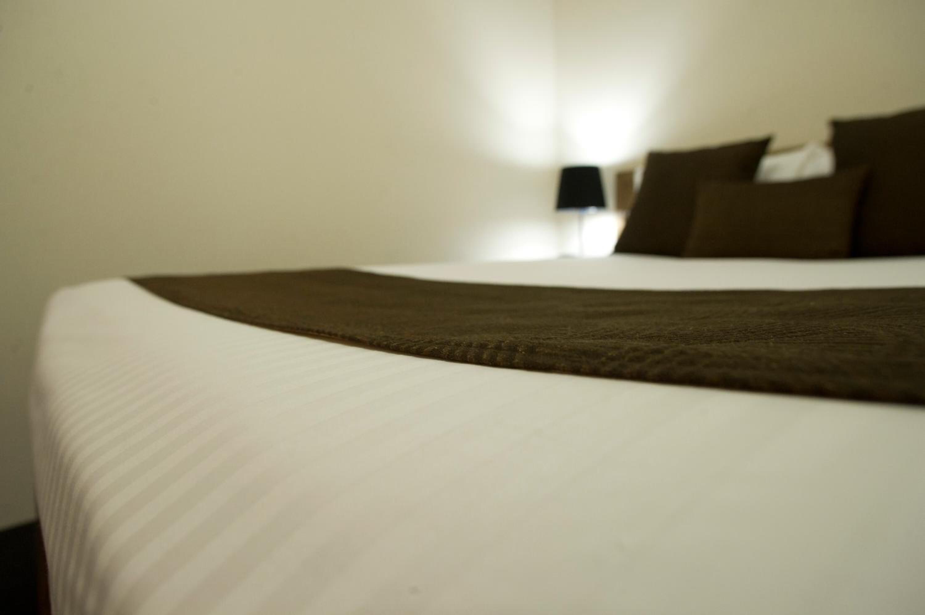 A neatly made bed with a white and brown bedspread, a black lamp on a bedside table, and a white wall in the background.