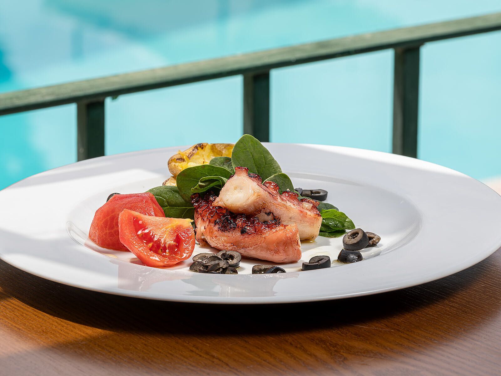 Prato branco com frango grelhado, tomates fatiados, espinafre e azeitonas pretas sobre uma mesa de madeira, com um corrimão azul ao fundo.
