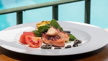 Prato branco com frango grelhado, tomates fatiados, espinafre e azeitonas pretas sobre uma mesa de madeira, com um corrimão azul ao fundo.