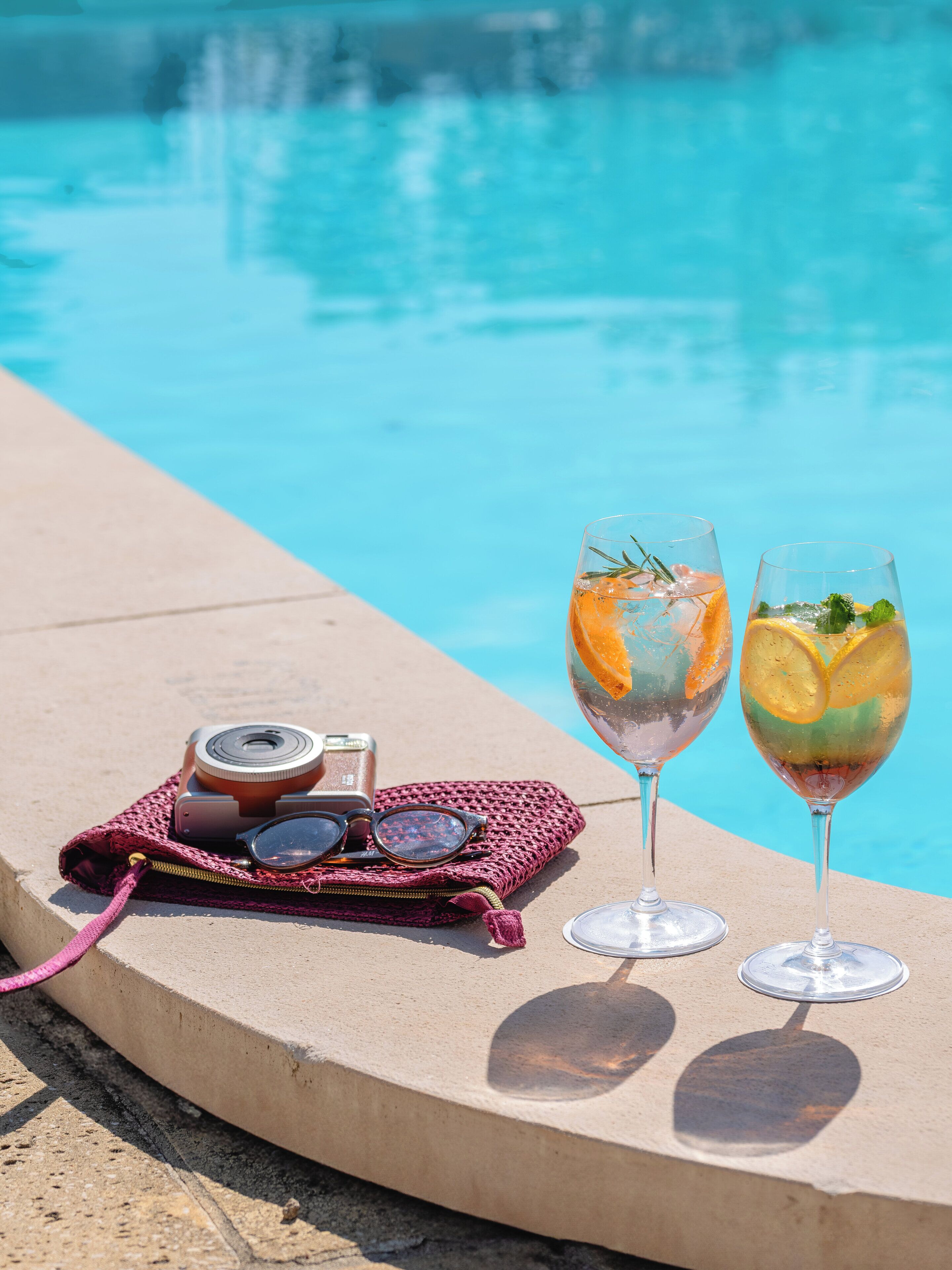 A poolside setting with two glasses of a citrus drink, a camera, and a pink bag.