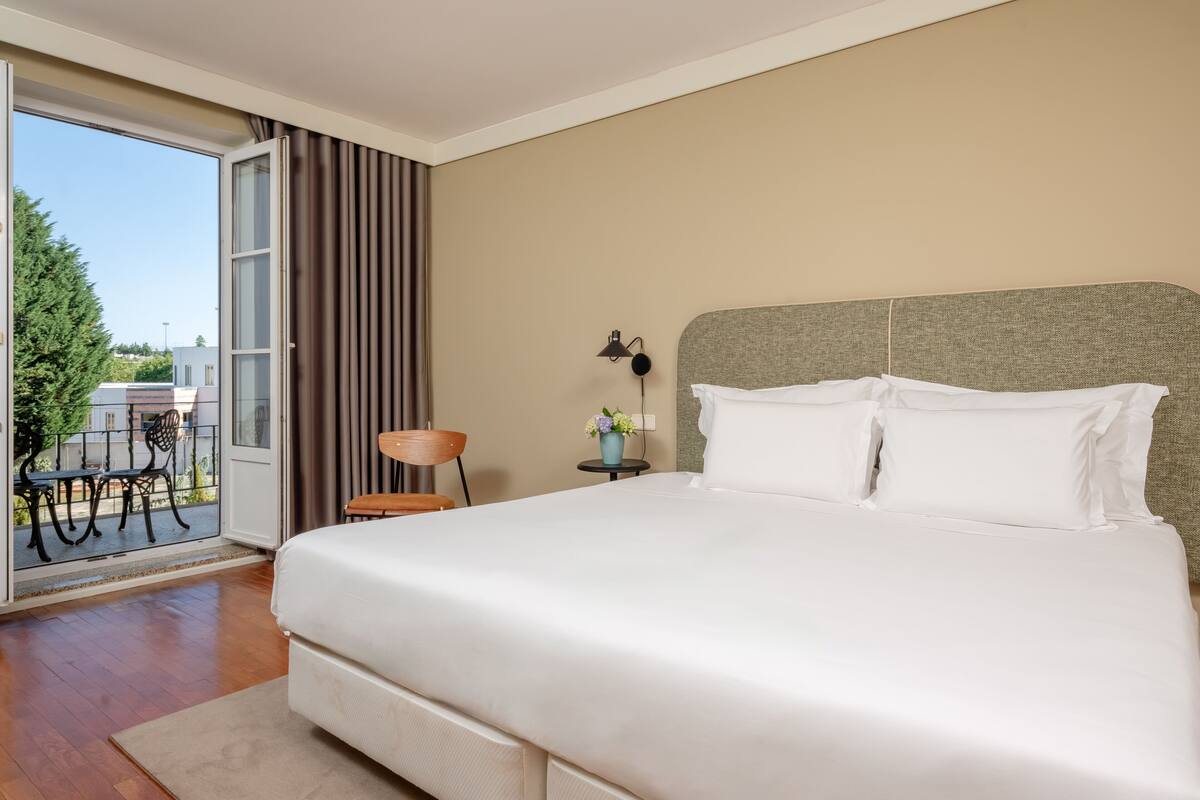 A hotel room with a large bed, a small table with a lamp, a chair, and a view of the outdoors.