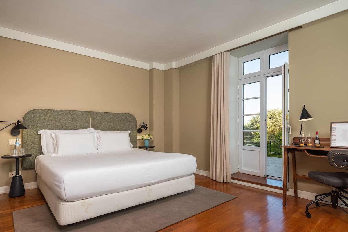 A hotel room with a large bed, a desk with a chair, a small table with a lamp, and a window with curtains.