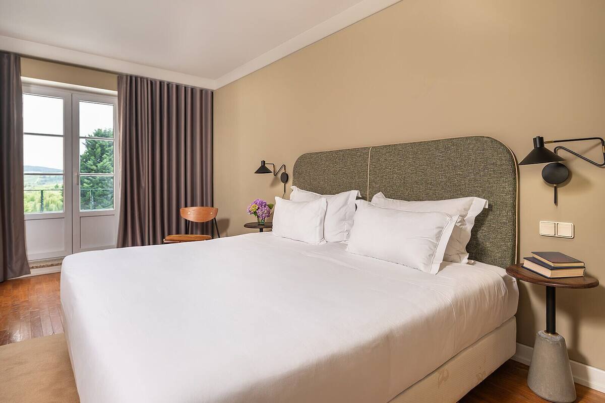 A large bed with white linens, a headboard with a textured pattern, a bedside table with a lamp, and a view of greenery through a window.