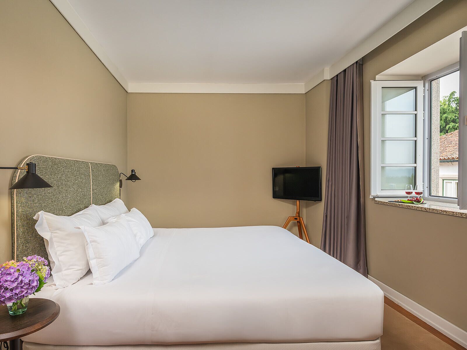 Quarto de hotel com uma cama grande, uma mesa de cabeceira com abajur, televisão e janela com cortinas.