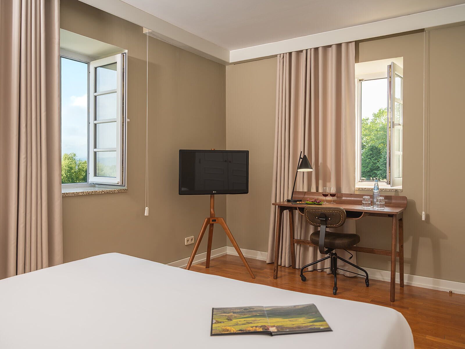 A hotel room with a bed, a desk with a computer, a TV on a stand, and a window with curtains.