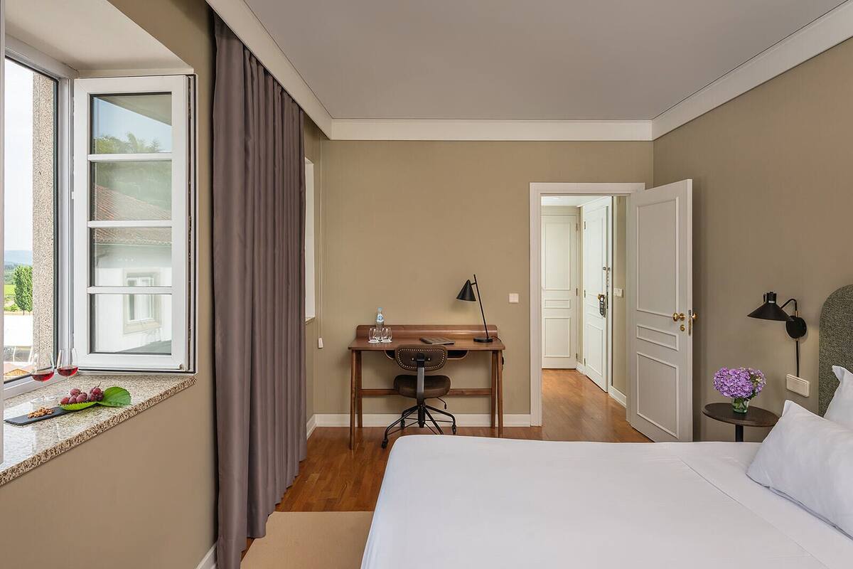 A hotel room with a bed, a desk with a chair, a window with curtains, a door, and a small table with a lamp.