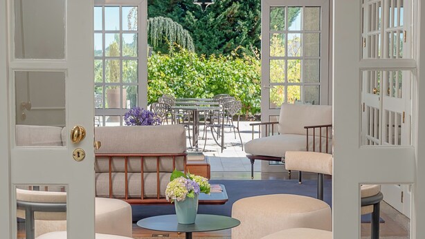 Une porte ouverte donne sur une pièce bien éclairée, dotée d’un plancher en bois, d’une table ronde entourée de chaises et d’un canapé.
