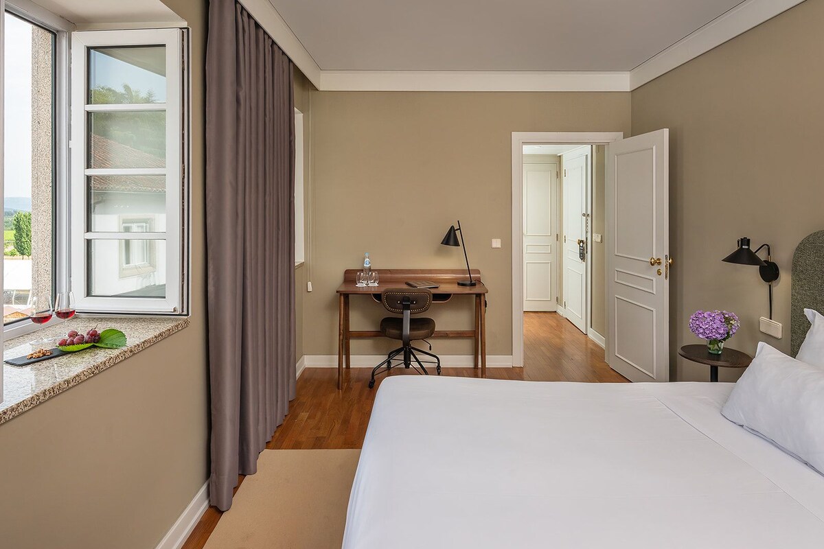 A hotel room with a bed, a desk with a chair, a window with curtains, a door, and a small table with a lamp.