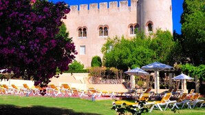 Un edificio de estilo castillo con una torre, rodeado de vegetación y una zona de piscina con tumbonas.