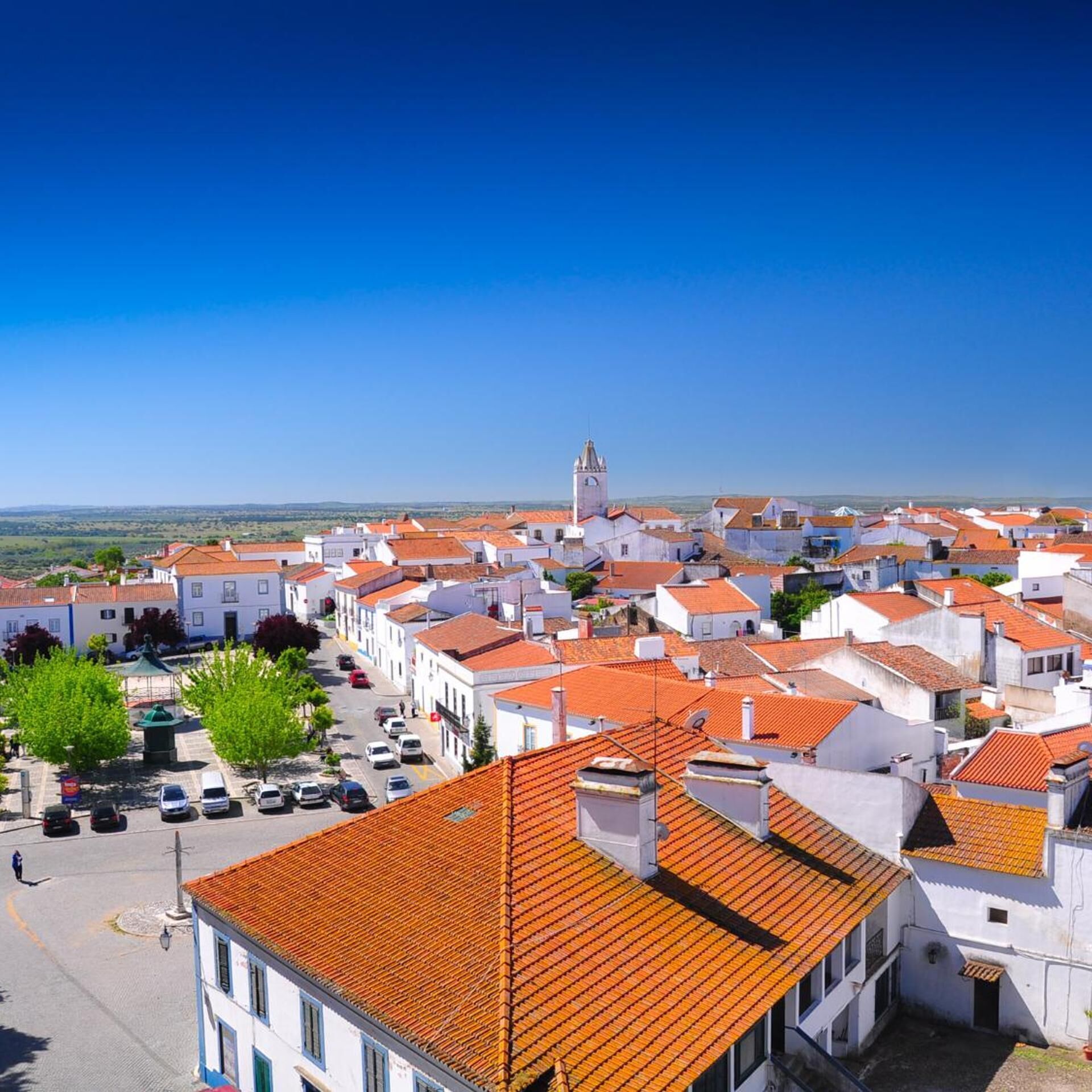 A town with white buildings and red roofs, a clear sky, and a distant view of the sea.