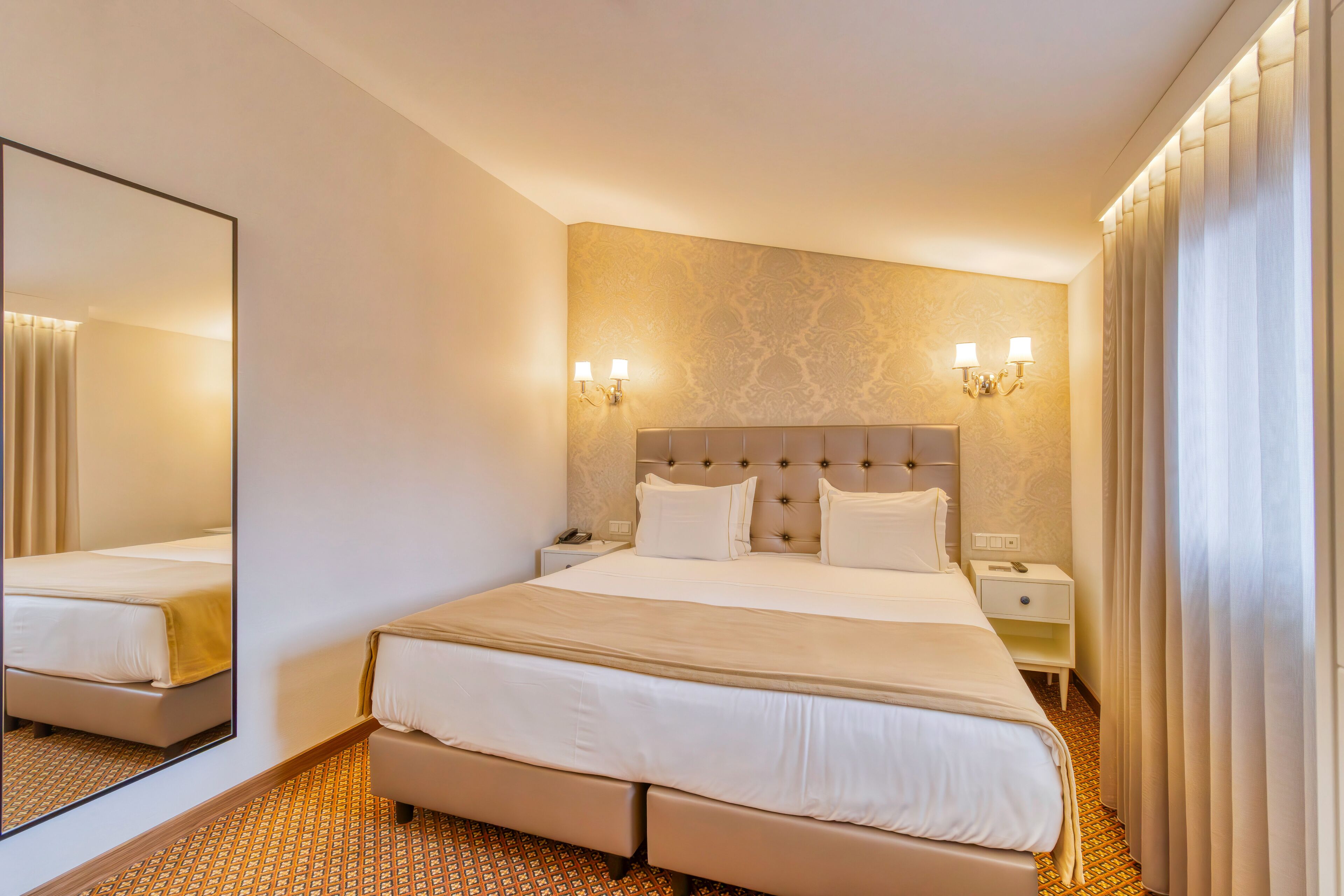 A hotel room with a large bed, two bedside tables, a nightstand, a mirror, and a wall-mounted light fixture.