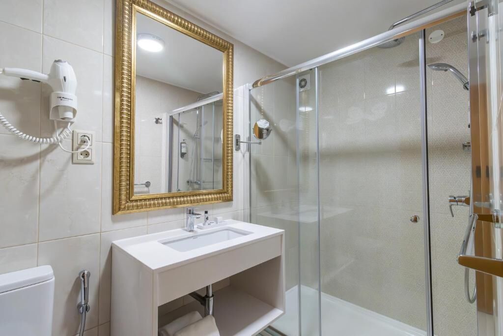 Un baño moderno con un lavamanos blanco, un espejo con marco dorado y una ducha con cerramiento de vidrio.