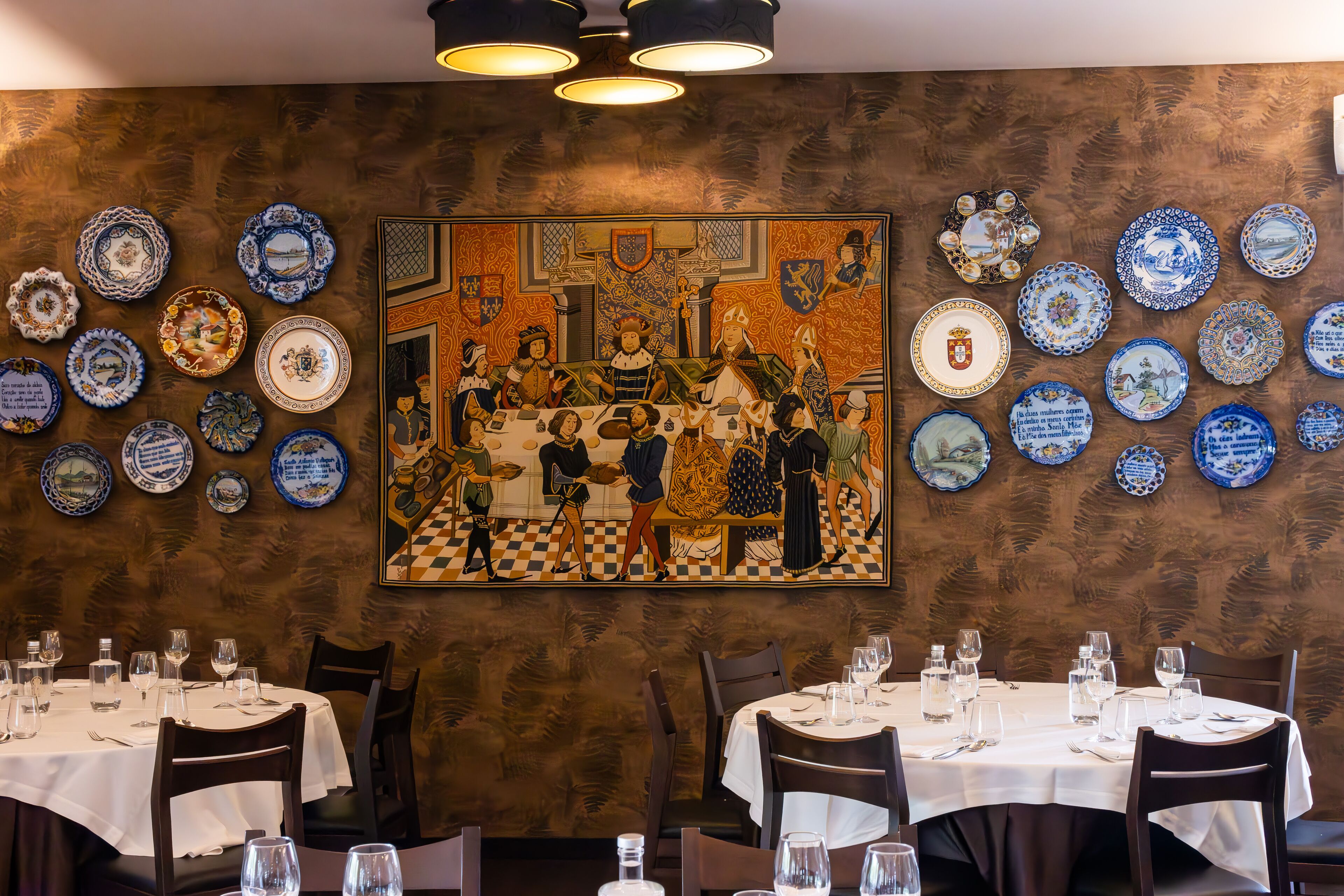 A dining area with round tables set for two, each with a white tablecloth, wine glasses, and cutlery. The walls are adorned with a large, detailed painting and numerous blue and white plates.