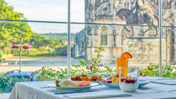 A table with breakfast items and a glass of orange juice, with a view of a historic building through a window. - Hotel Lis Batalha (Batalha)