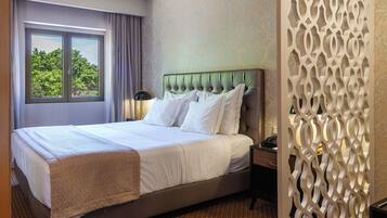 A hotel room with a large bed, a window with curtains, a decorative wall panel, and a nightstand with a lamp.