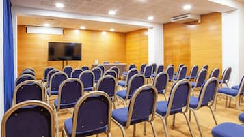 Un salón de conferencias con filas de sillas azules, un televisor de pantalla plana grande, paneles de madera en las paredes y suelo alfombrado.