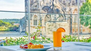 A table with a plate of food and a glass of drink, with a view of a historic building through a window.