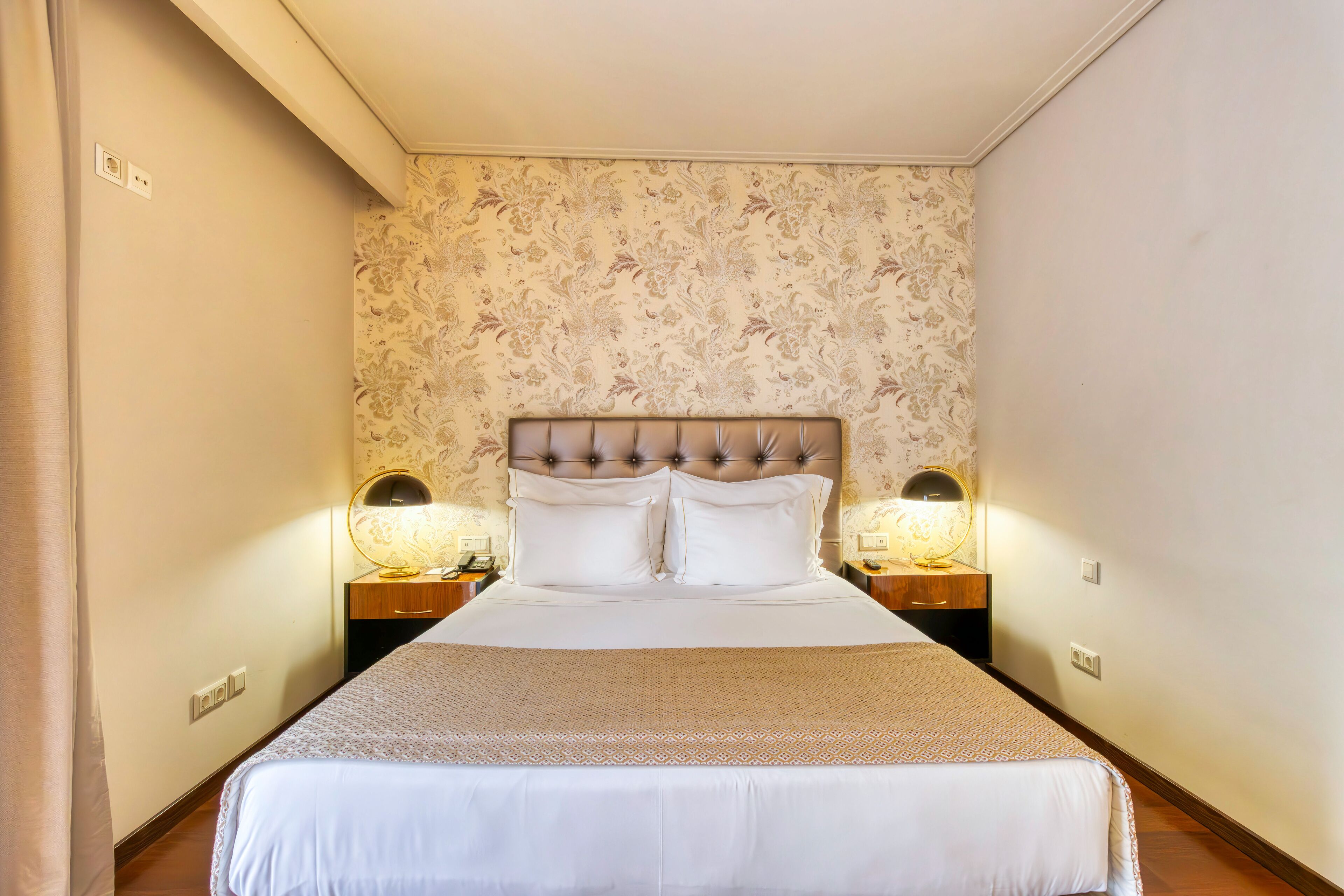 A neatly made bed with a headboard, two bedside lamps, and a patterned wallpaper.