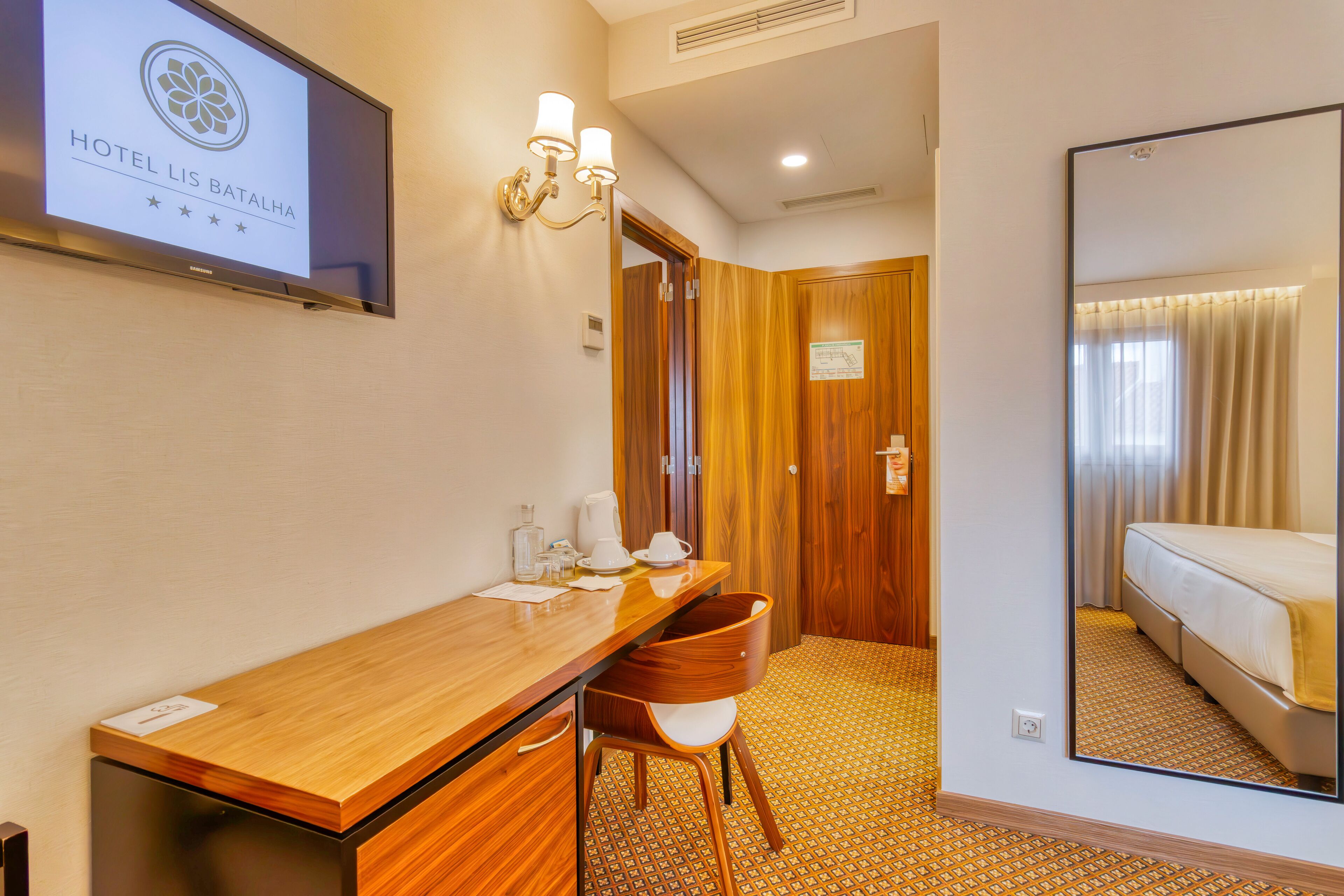 A hotel room with a wooden desk, a chair, a bed, and a large mirror.