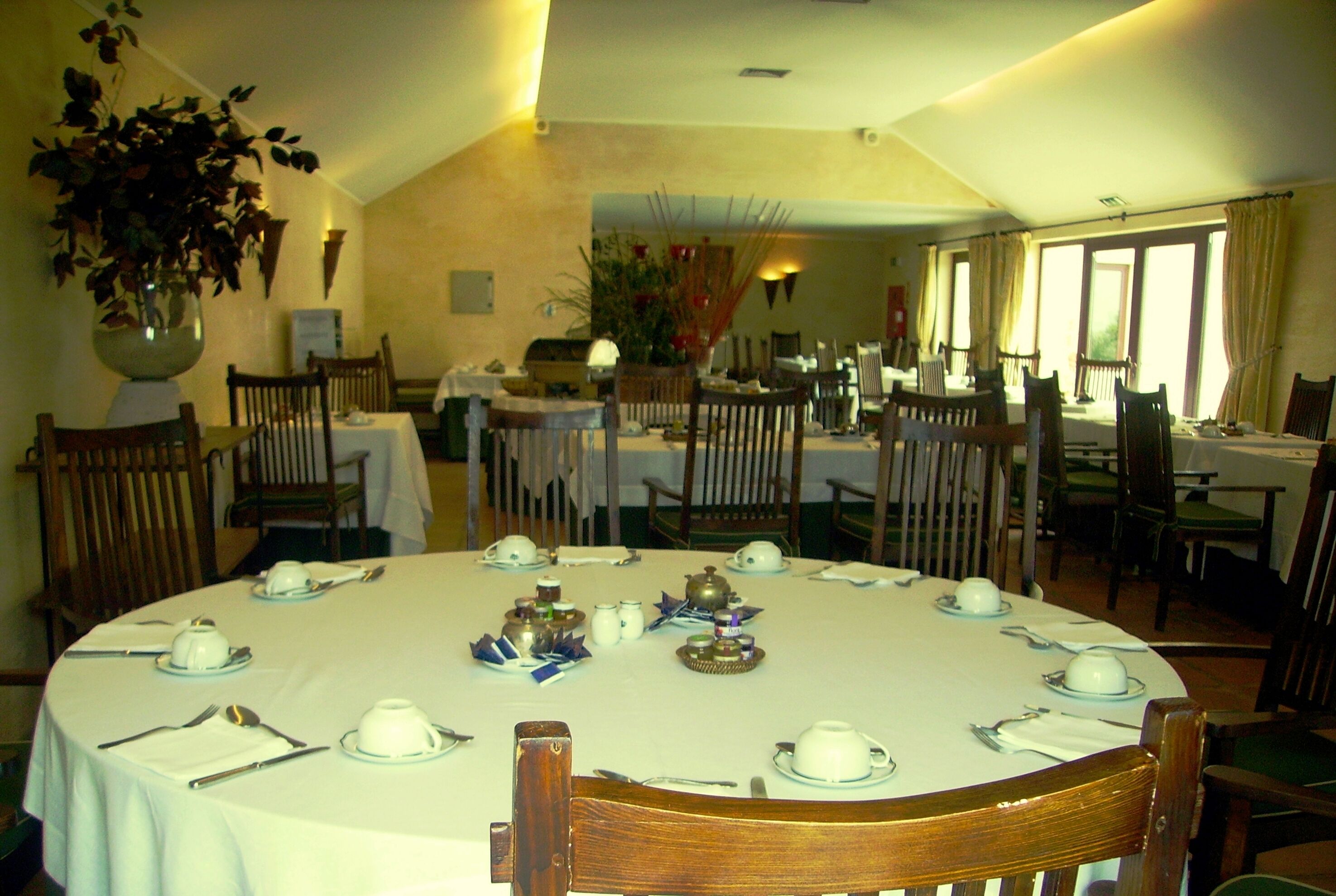 Comedor con mesas redondas dispuestas para una comida, sillas y un jarrón grande de flores.