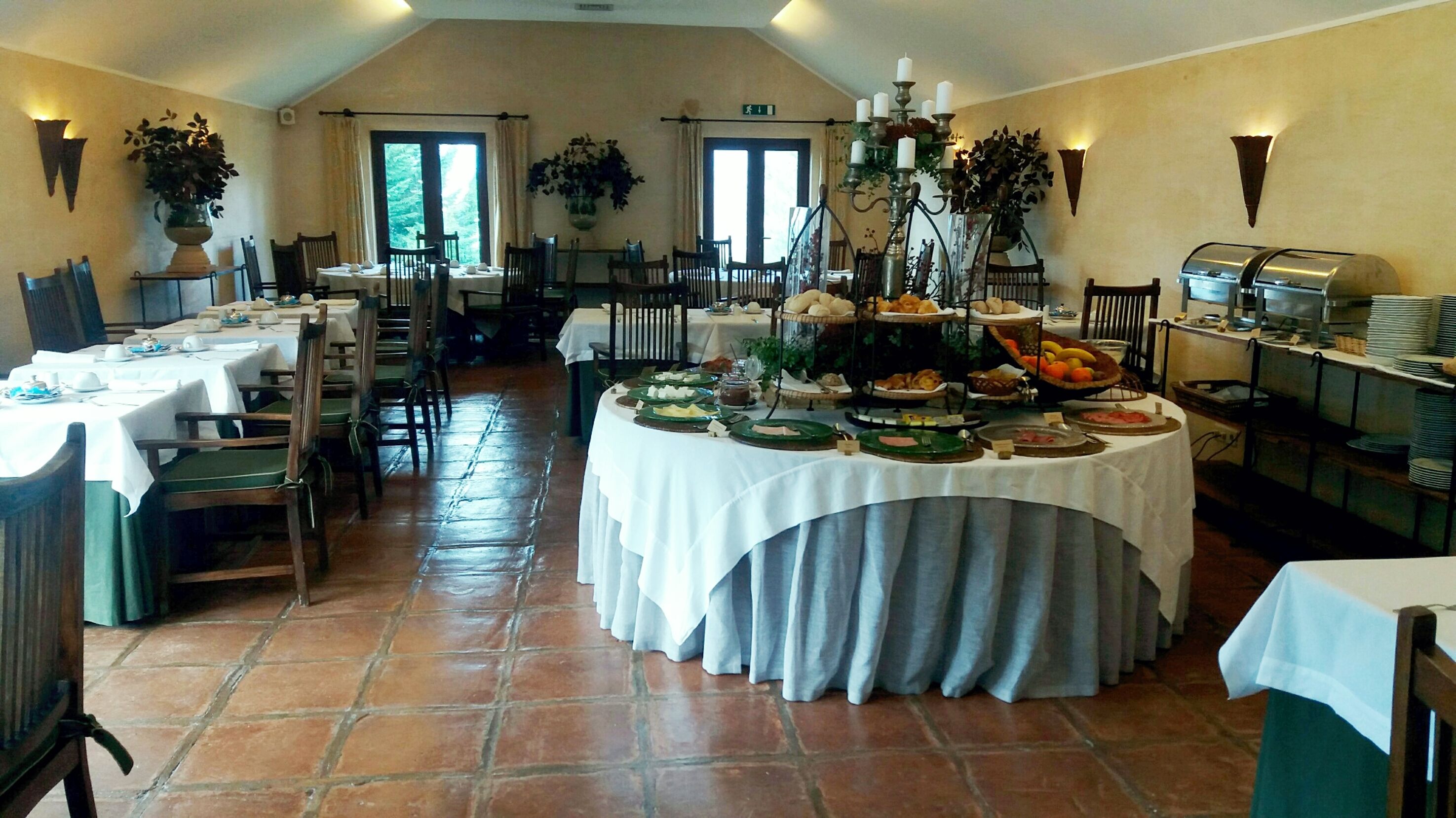 A dining area with multiple tables set for breakfast, featuring a variety of food items and a buffet setup.