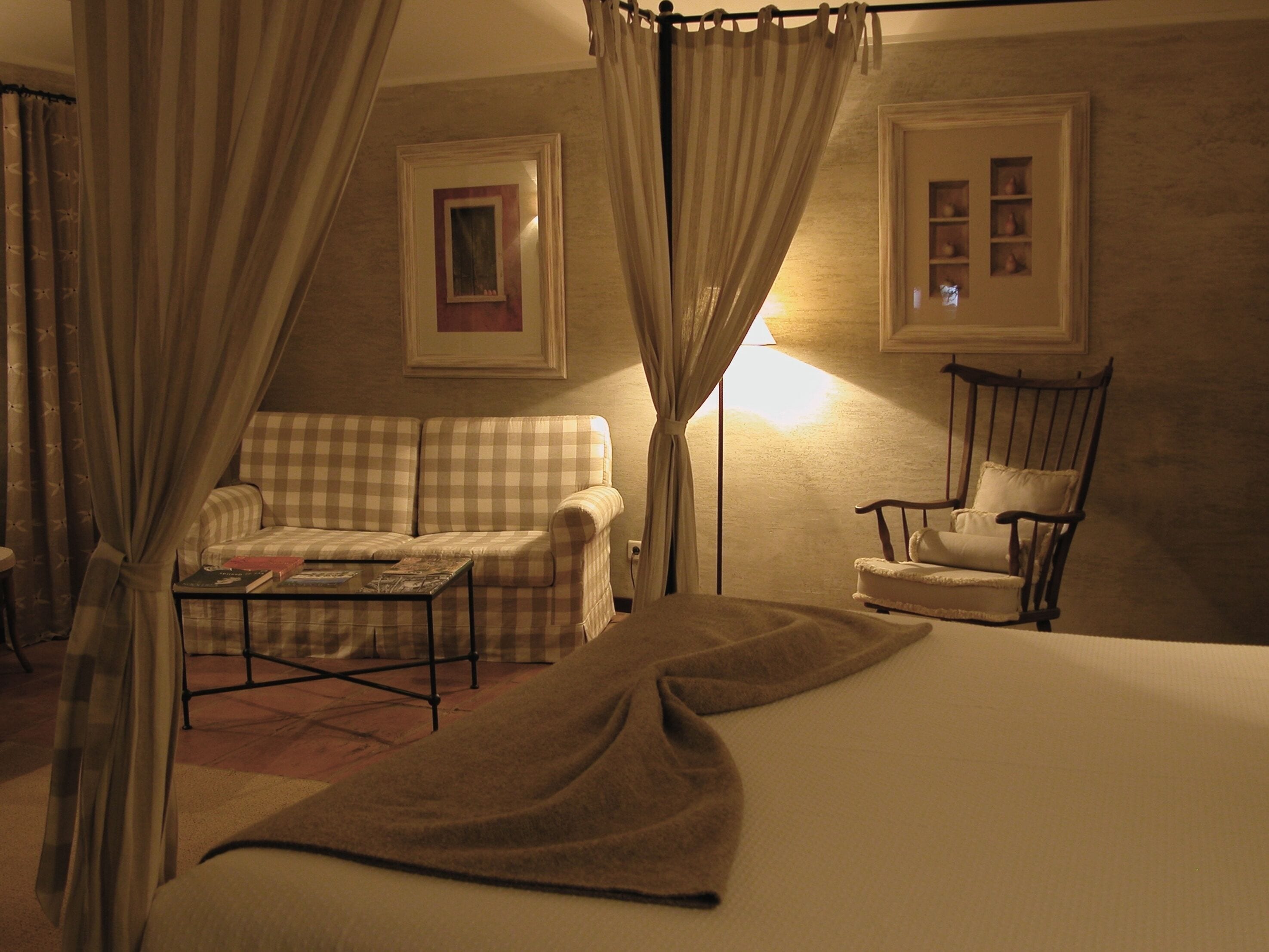 A bedroom with a bed, a checkered sofa, a chair, a small table, and curtains.