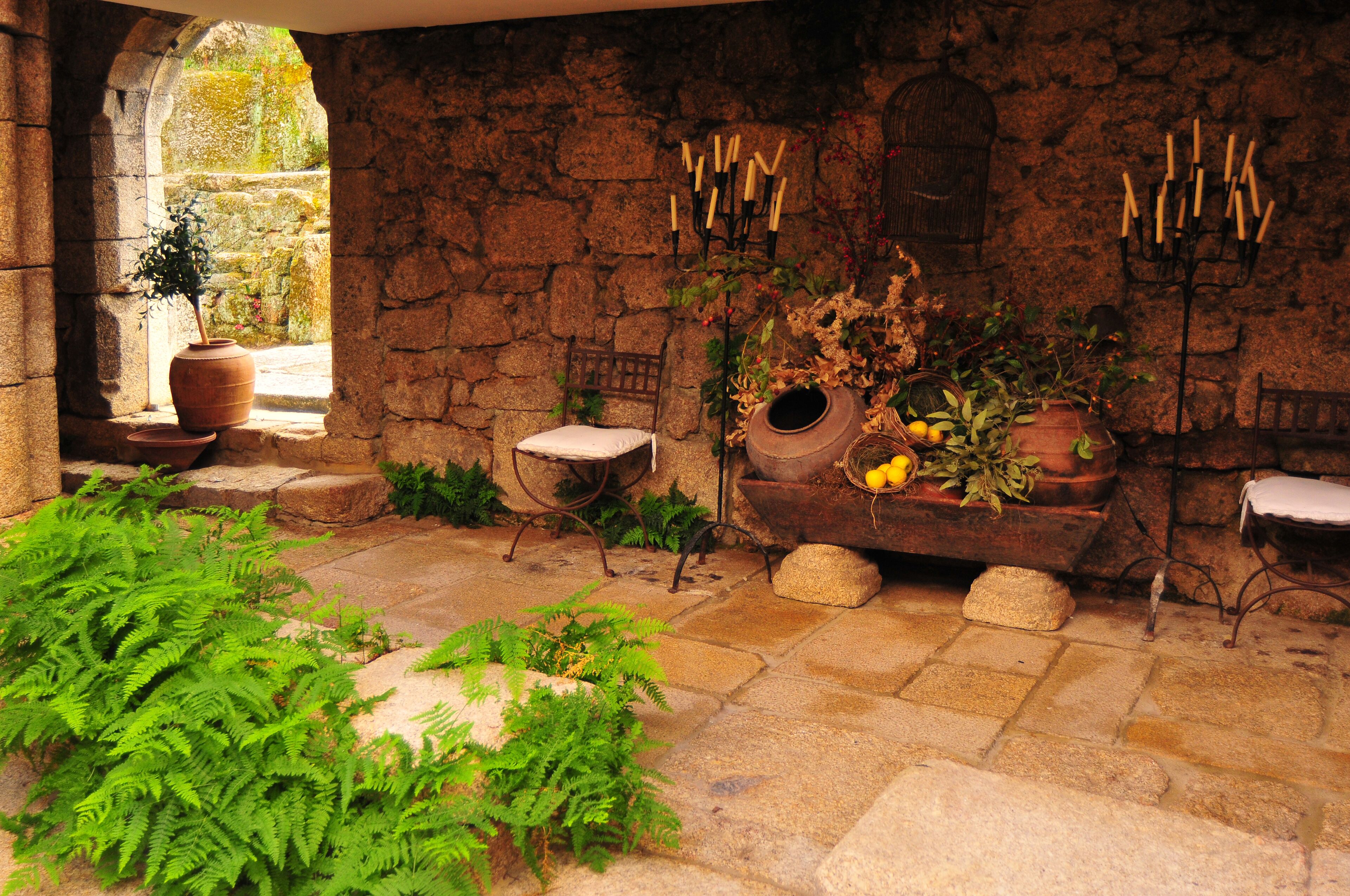Una habitación de paredes de piedra con decoración rústica, que incluye una ventana, una planta en maceta, una silla de madera y una mesa con un florero.