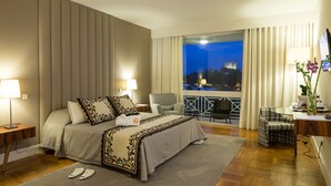 A hotel room with a large bed, a desk, a chair, and a view of a castle at night. - Pousada de Bragança - São Bartolomeu (Braganca)
